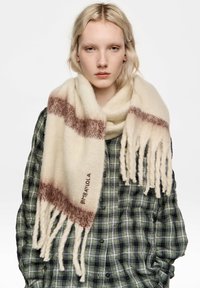 Kremowy szal z brązowymi paskami, wykończony frędzlami, o miękkiej fakturze; posiada haftowane logo. Noszony na zielonej koszuli w kratę.