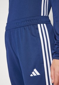 Navy blue athletic tracksuit with white side stripes, featuring a textured fabric, elastic waistband, and the Adidas logo in white on the leg.
