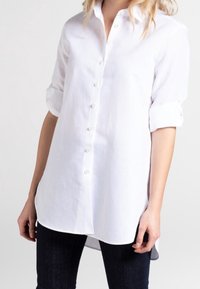 Camicia bianca con bottoni, realizzata in tessuto leggero, con colletto, maniche arrotolate e orlo curvo. Presenta bottoni simili a perle lungo la parte anteriore.
