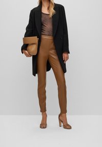 Svart kappa över en bronzfärgad topp, tillsammans med bruna läderbyxor och bruna klacksandaler. Bär en tan geometrisk clutchtvätt. Släta texturer.