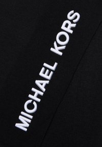Juoda medžiaga su tekstūruotu mezgimo raštu; vienoje pusėje yra balta siuvinėta užrašas "MICHAEL KORS".