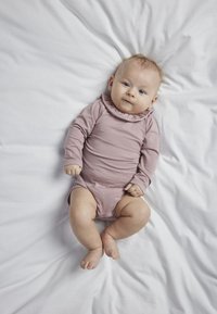 Bodysuit za bebe dugih rukava od mekane, prašnjavo ružičaste tkanine s volanima na izrezu, leži na naboranoj bijeloj površini.