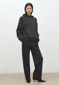 Felpa nera oversize con tasca a marsupio, abbinata a pantaloni neri a gamba larga e tacchi neri a punta su sfondo chiaro.