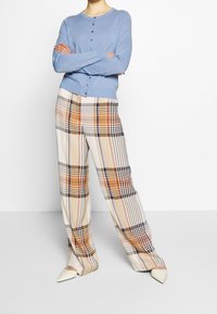 Person, die einen blauen Knopfcardigan, weit geschnittene karierte Hose in Beige, Orange und Marineblau sowie weiße spitze Schuhe trägt, steht mit verschränkten Armen.