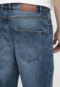 Zilas džinsa bikses ar teksturētu apdari, ādas jostasvietas uzlika ar uzrakstu "ISOLID" un divām muguras kabatām ar izliektiem stūriem.