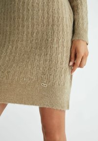 Abito maglione a coste beige con un motivo testurizzato e un logo del marchio in oro sul lato inferiore. Materiale morbido e caldo.