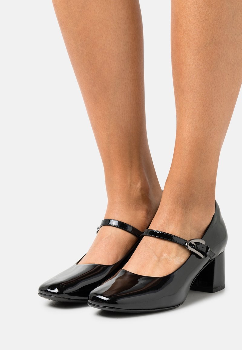 Jambes portant des chaussures Mary Jane en cuir verni noir, à bouts arrondis, brides à la cheville et talons blocs bas sur fond clair.