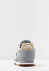 Zapatilla New Balance en tonos gris y beige vista desde atrás, mostrando una tela texturizada y una suela de goma sobre fondo blanco.