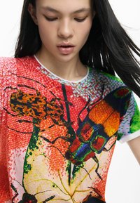 Színes póló élénk narancssárga, zöld és piros rovarmintával. Kerek nyakkivágás fehér szegéllyel; rövid ujjú. Texturált anyag.