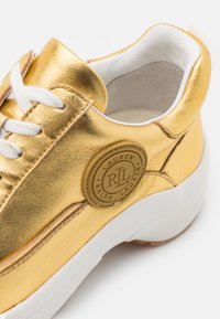 Zlaté metalické tenisky s bílými tkaničkami, polstrovaným bílým interiérem, bílou podešví a kulatým logem "Lauren Ralph Lauren" na boku.