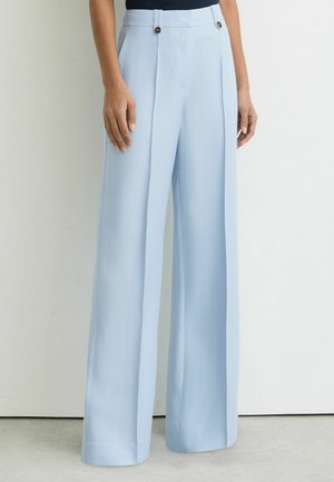 Femeie purtând pantaloni largi, cu talie înaltă, de culoare bleu deschis, cu detalii cu nasturi lângă talie, stând în picioare lângă un perete simplu.