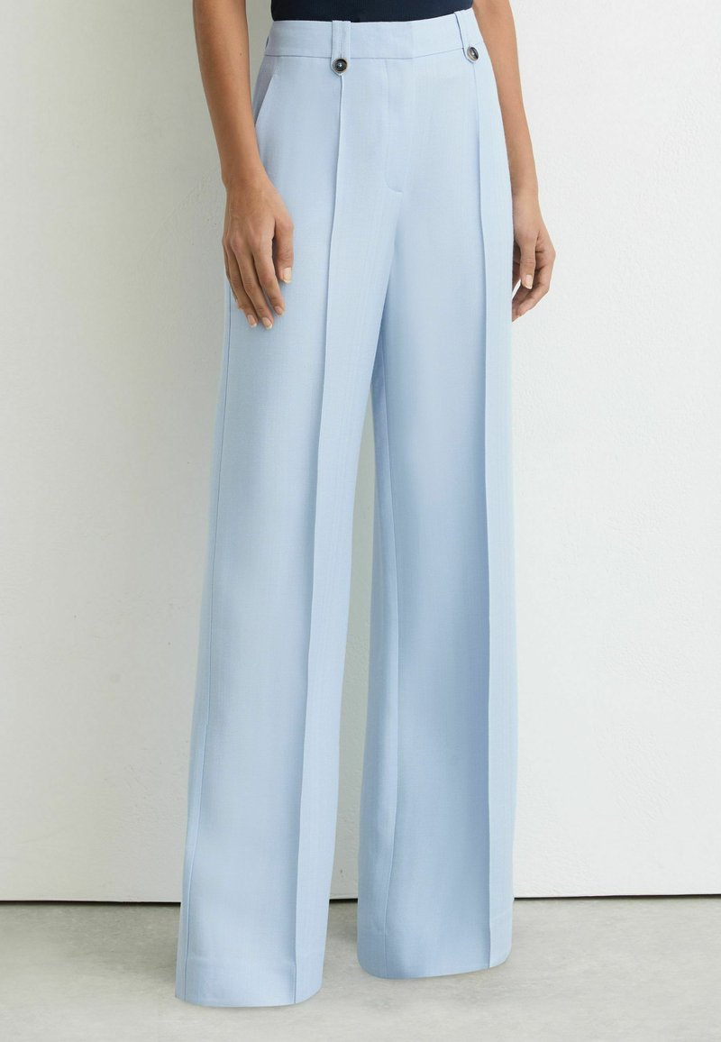 Žena stojaca pri holom múre, oblečená vo vysokých svetlomodrých nohaviciach s širokými nohavicami a gombíkmi pri páse.