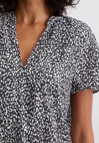 Femme portant un chemisier à manches courtes noir et blanc à pois, avec un col froncé et une ouverture en forme de V.