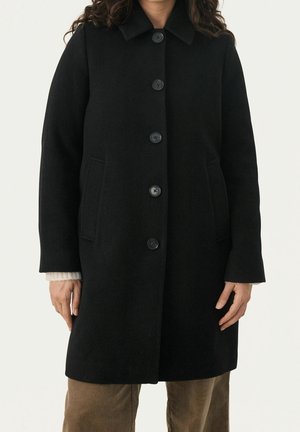 Manteau en laine noire à cinq boutons, col cranté et poches latérales porté sur un pull beige et un pantalon marron.