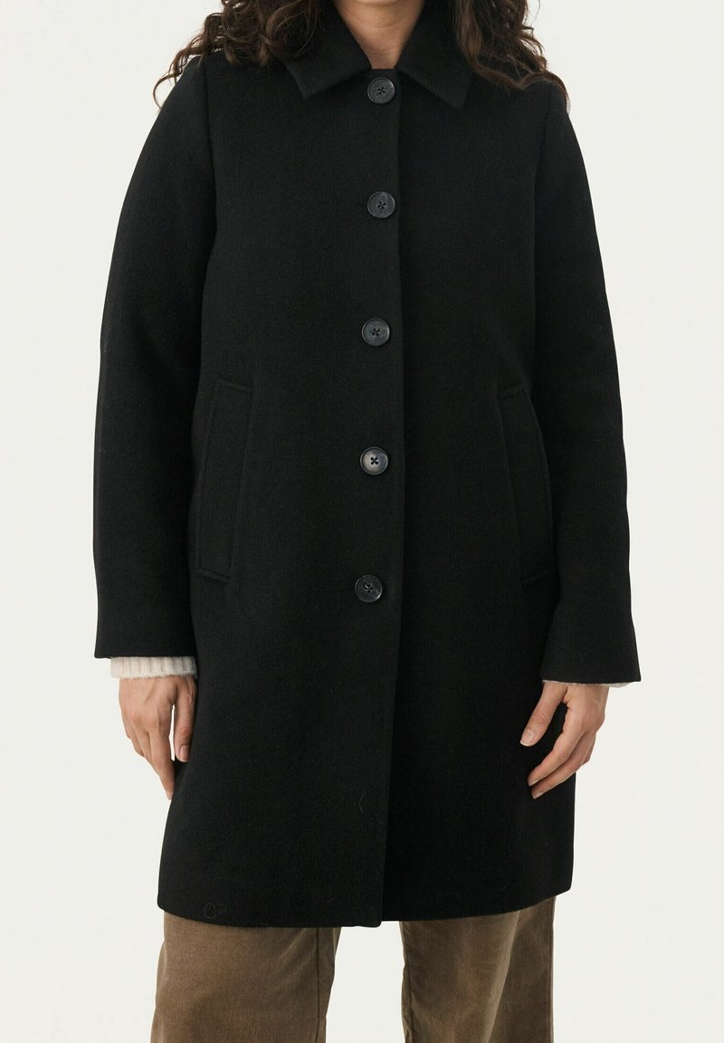 Manteau en laine noire à cinq boutons, col cranté et poches latérales porté sur un pull beige et un pantalon marron.