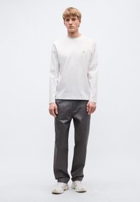 Fehér hosszú ujjú ing logóval a mellkason, szürke bő fazonú nadrággal és világos színű sportcipővel párosítva. Egyszerű, modern kialakítás.