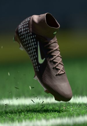 Καφέ ποδοσφαιρικό παπούτσι Nike με λευκό σχέδιο swoosh, με καρφιά στη σόλα, σηκώνοντας χορτάρι σε σημαδεμένο πράσινο γήπεδο κάτω από χαμηλό φωτισμό.