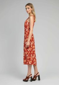 Robe midi florale rouge avec des bretelles spaghetti, présentant un motif botanique orange. Associée à des sandales à talons marron, présentée sur un fond neutre.