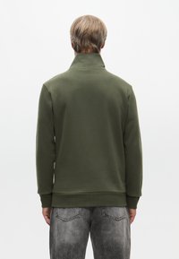 Zöld polár pulóver magas gallérral, hosszú ujjakkal és bordás aljjal. Világosszürke, elhalványult farmernadrággal viselve. Egyszerű dizájn, egyszínű.