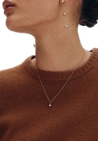 Złote kolczyki z akcentami w kształcie gwiazdy oraz pasujący złoty naszyjnik z małym wisiorkiem w kształcie gwiazdy na modelce w brązowym swetrze.