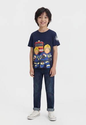 Niño de pie sonriendo con una camiseta azul marino con dibujos animados de bomberos y camión de bomberos, jeans oscuros y zapatillas blancas sobre fondo blanco.