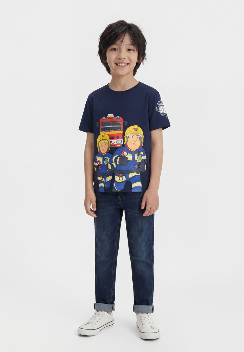Αγόρι που στέκεται και χαμογελά φορώντας μπλε σκούρο μπλουζάκι με κινούμενους ήρωες πυροσβεστών και πυροσβεστικό όχημα, σκούρο τζιν και λευκά αθλητικά σε λευκό φόντο.