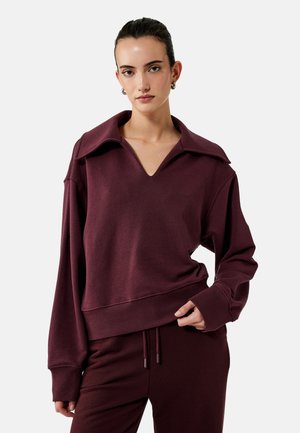 Vrouw draagt een bordeaux oversized sweatshirt met brede kraag en bijpassende joggers, staand tegen een eenvoudige lichte achtergrond.