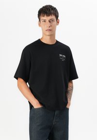 Sort bomulds T-shirt med rund halsudskæring og korte ærmer. Har minimal hvid tekst på brystet. Stylet med mørke denim jeans.