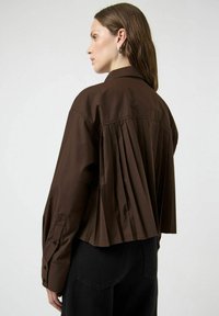 Camicia marrone con bottoni, dettaglio a pieghe sulla schiena e maniche lunghe, realizzata in tessuto liscio, con colletto classico e accenti neri.