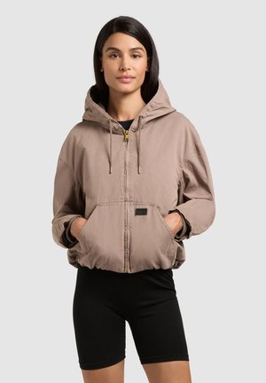 Young woman wearing a tan zip-up hooded jacket with hands in front pockets and black fitted shorts against plain background.