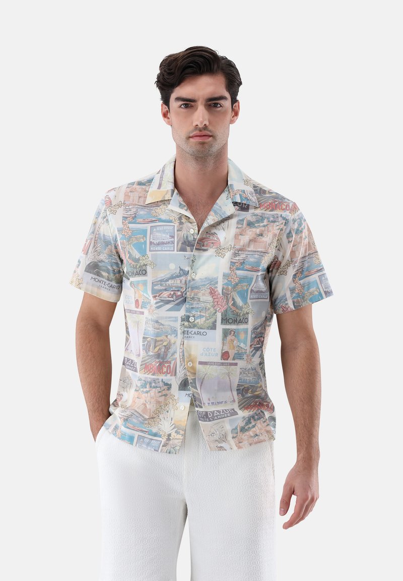 Mann trägt ein kurzärmeliges Hemd mit Vintage-Monaco- und Côte d'Azur-Reiseposter-Drucken sowie weiße strukturierte Hosen.