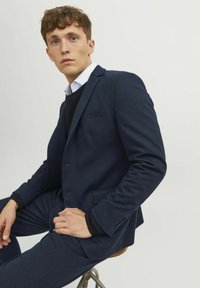 Blazer blu navy su misura con vestibilità slim, rever a punta, tasca frontale singola e polsini con bottone. Indossato sopra un maglione blu navy e una camicia bianca.