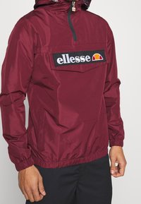 Red windbreaker jacket with a half-zip, elastic cuffs, and a front pocket featuring an embroidered logo on a black patch.