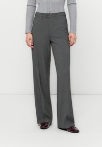 Pantaloni grigi, a gamba larga e su misura, con una texture liscia, chiusura con bottone frontale e due tasche laterali, abbinati a scarpe scure.