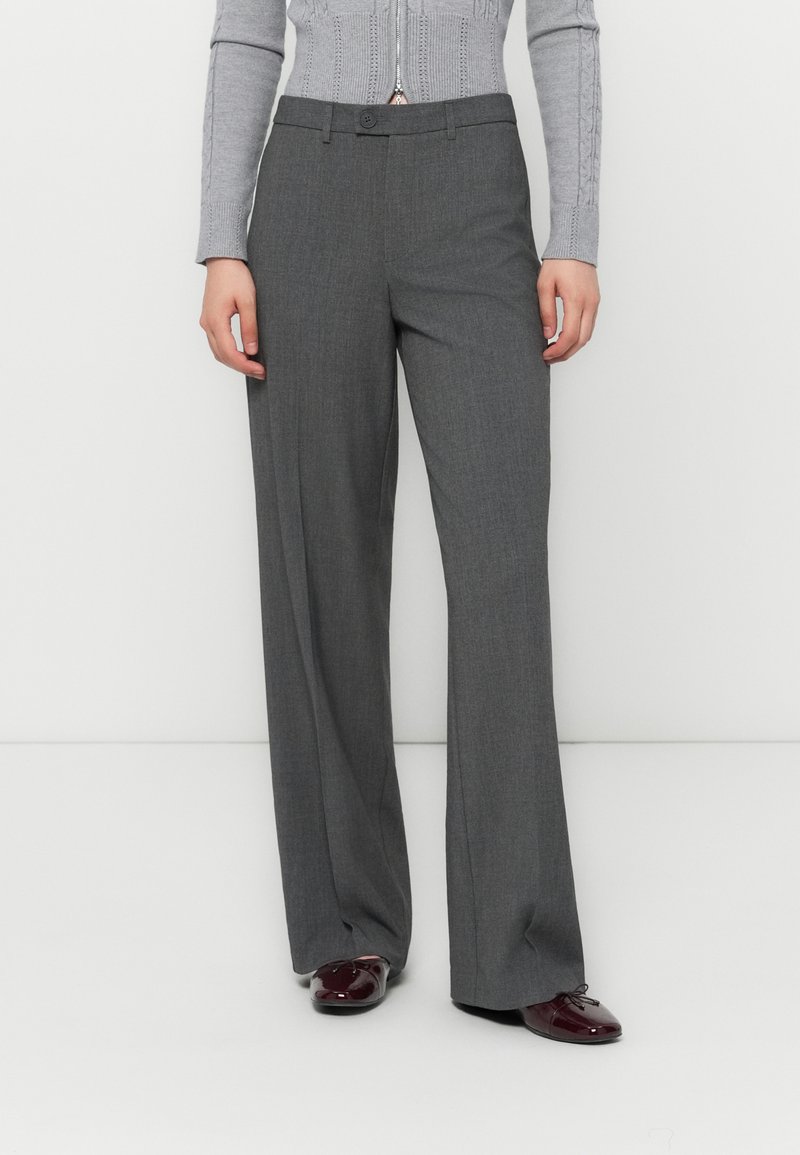 Pantaloni grigi, a gamba larga e su misura, con una texture liscia, chiusura con bottone frontale e due tasche laterali, abbinati a scarpe scure.