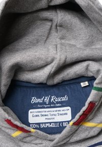 Sudadera de algodón gris con una textura suave, que presenta una etiqueta interior de color azul marino. Los acentos incluyen franjas rojas, amarillas y verdes cerca del collar.