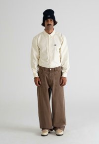 Light beige button-up shirt with an embroidered logo, paired with loose brown pants and beige shoes. The model wears a dark beanie.
