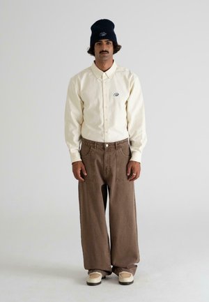 Light beige button-up shirt with an embroidered logo, paired with loose brown pants and beige shoes. The model wears a dark beanie.