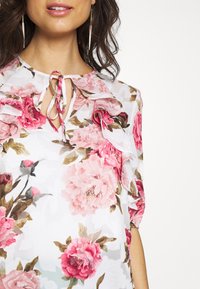 Květinová blůza s růžovými a bílými květy, průhlednou texturou, volánkovými rukávy, kulatým výstřihem a detaily na zavázání vpředu.