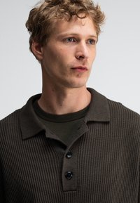 Braunes geripptes Strickpolo mit einer Knopfleiste mit drei Knöpfen und einem spitzen Kragen, getragen über einem dunkelgrünen Rundhals-Shirt.