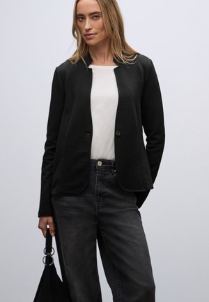 Zwarte gestructureerde blazer met een ronde kraag, sluiting met één knoop en twee zakken aan de voorkant. Gepaard met een witte top en grijze spijkerbroek.