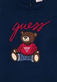 Tengerészkék pulóver, amelyen egy piros felsőt és kék farmert viselő maci hímzett, a grafika felett pedig piros, kurzív "Guess" felirat található.
