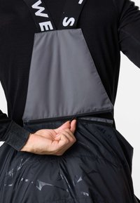 Persona che indossa salopette nera e grigia con bretelle brandizzate, che allaccia o slaccia una cerniera sulla schiena all'altezza della vita.