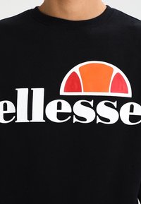 Sudadera negra con el logotipo blanco de "ellesse" y un gráfico redondo colorido en naranja y rojo encima del texto. Tejido suave y texturizado.