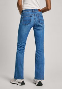 Jeans acampanados de mezclilla azul con una cintura de altura media, dos bolsillos traseros con detalles de costura y un sutil parche de cuero en la cintura.