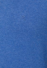 Tissu tricoté bleu avec un petit logo brodé représentant des ciseaux croisés et un bouton cousu avec un fil bleu assorti.