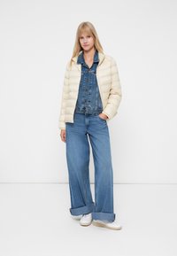 Jasno beżowa pikowana kurtka na dżinsowym kombinezonie i szerokich niebieskich dżinsach, uzupełniona białymi sneakersami. Gładka tekstura, casualowy design.