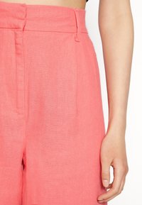 Korálové lněné kalhoty s vysokým pasem, předními sklady a bočními kapsami; lehká, texturovaná tkanina; čistý design bez zdobení.