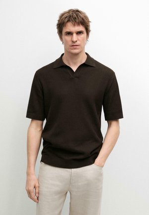 Mężczyzna ubrany w ciemnobrązową, prążkowaną koszulkę polo z krótkim rękawem oraz jasnobeżowe spodnie, stojący na jednolitym białym tle.