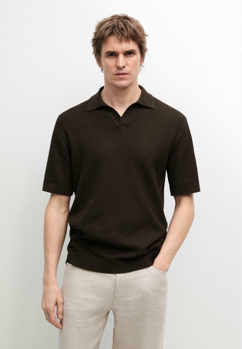 Mann trägt ein dunkelbraunes, kurzärmeliges, geripptes Polohemd und hellbeige Hosen, steht vor weißem Hintergrund.